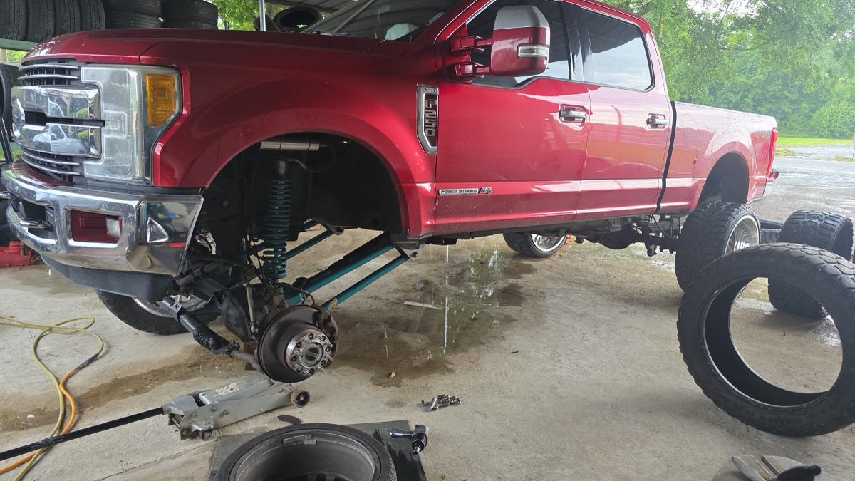 A lifted red Ford F-250 up on a jack in the shop with a wheel off, mid truck service